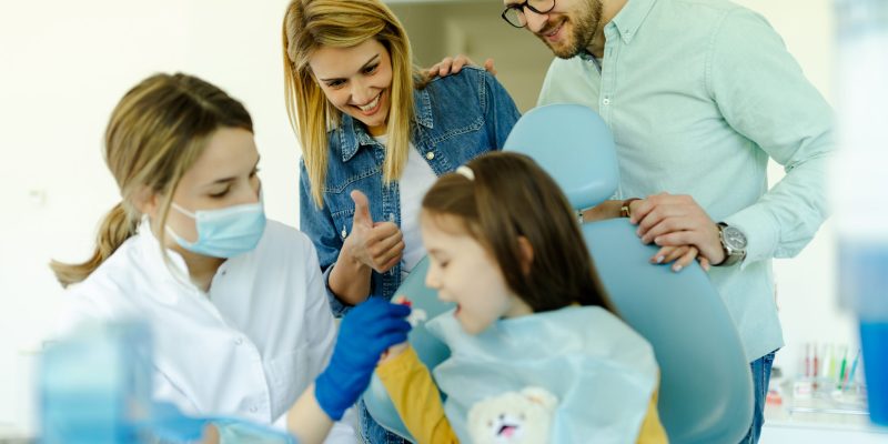 Dental professionals assisting a child patient at a bright dental clinic around a dental chair and chair-side assistants nearby to help during treatment。