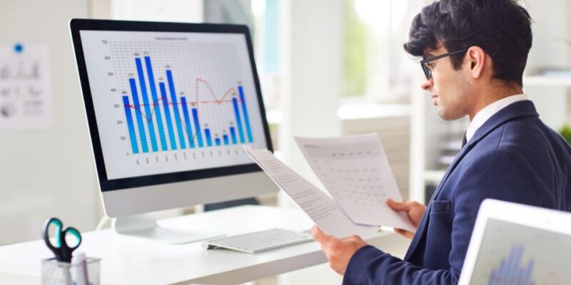 Businessperson in a suit reviews bar and line charts on a desktop monitor while holding documents in an office setting.