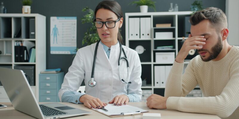 Female doctor in a white coat and stethoscope works at a desk while a man rests his head in his hand, suggesting a medical consultation.