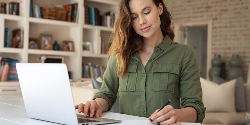 Woman in a green shirt types on a laptop at a white desk, writing in a notebook in a home office setting with shelves behind her.