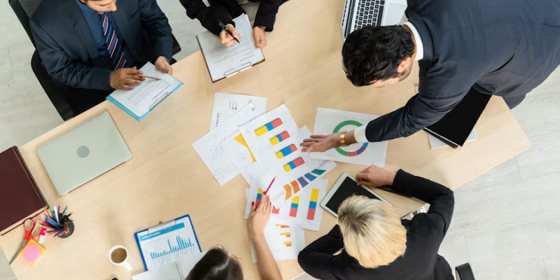 Overhead view of five professionals around a conference table examining colorful charts and graphs, with laptops and documents nearby.