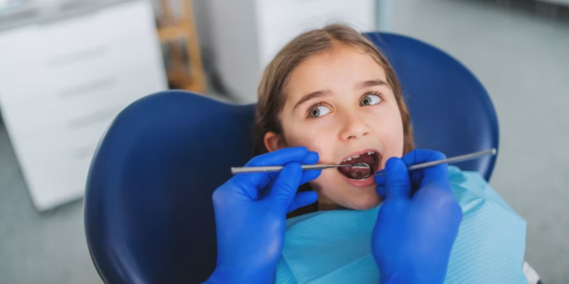 Dentist in blue gloves examines a young girl's mouth with dental tools in a chair.