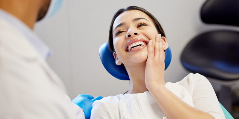 Smiling woman in a dental chair with a bib, interacting with a dentist wearing a mask and gloves.